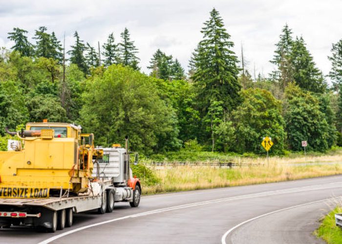 Powerful big rig semi truck with orange fenders ang oversize load sign on the bumper transporting heavy-duty tractor on the step down semi trailer running on the straight multiline highway road